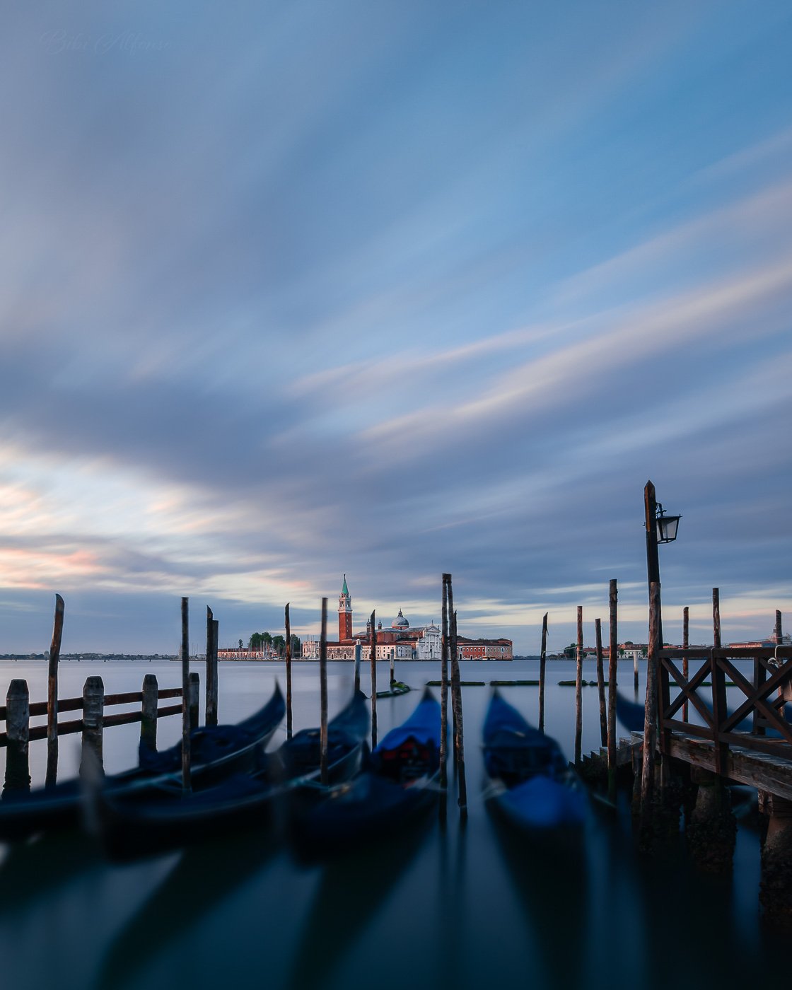 Long exposure morning scene in Venice, Italy, showing blurred gondolas in the foreground and the island of San Giorgio Maggiore in the distance under sweeping clouds. The image captures soft blue and golden tones, calm water, and gentle motion in the sky, evoking stillness and harmony between city and nature.