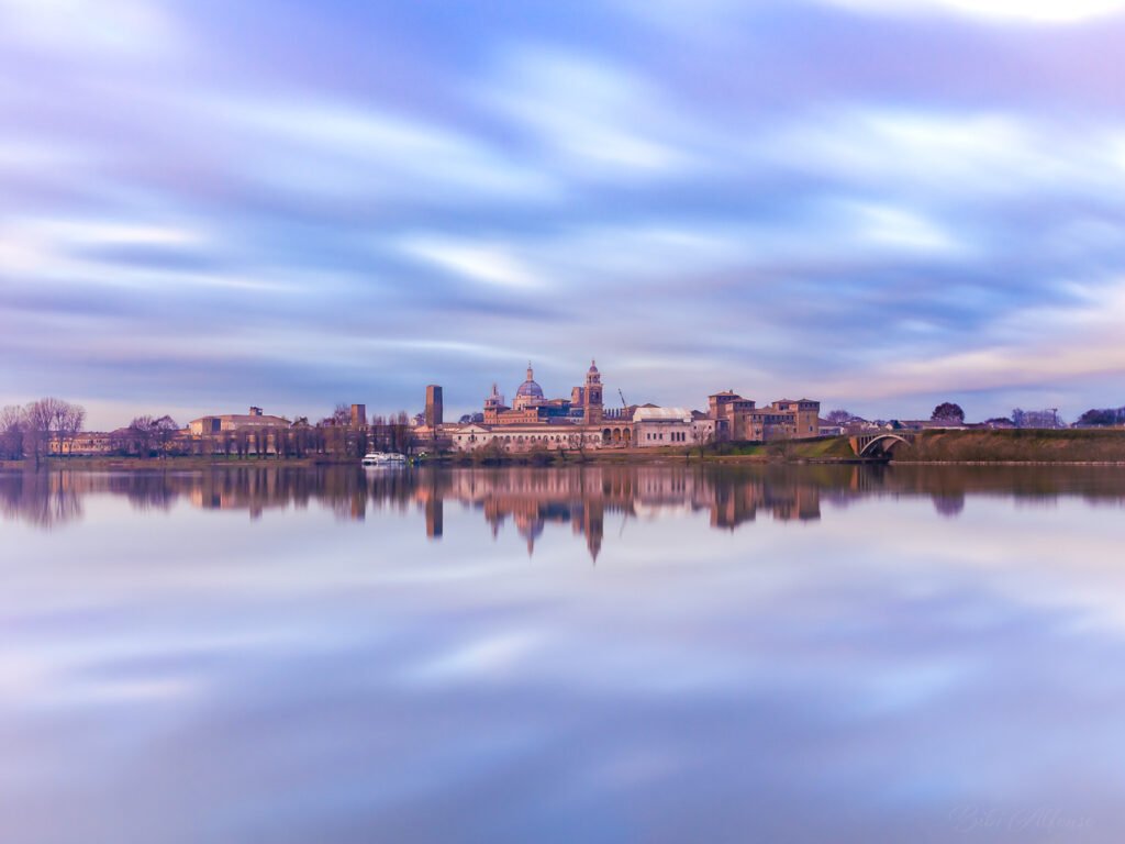 Spectacular long exposure of the Mantova skyline in the soft morning light, with streaky clouds above and the historic architecture reflected in the still water below, creating the effect of the city suspended in mid-air.
