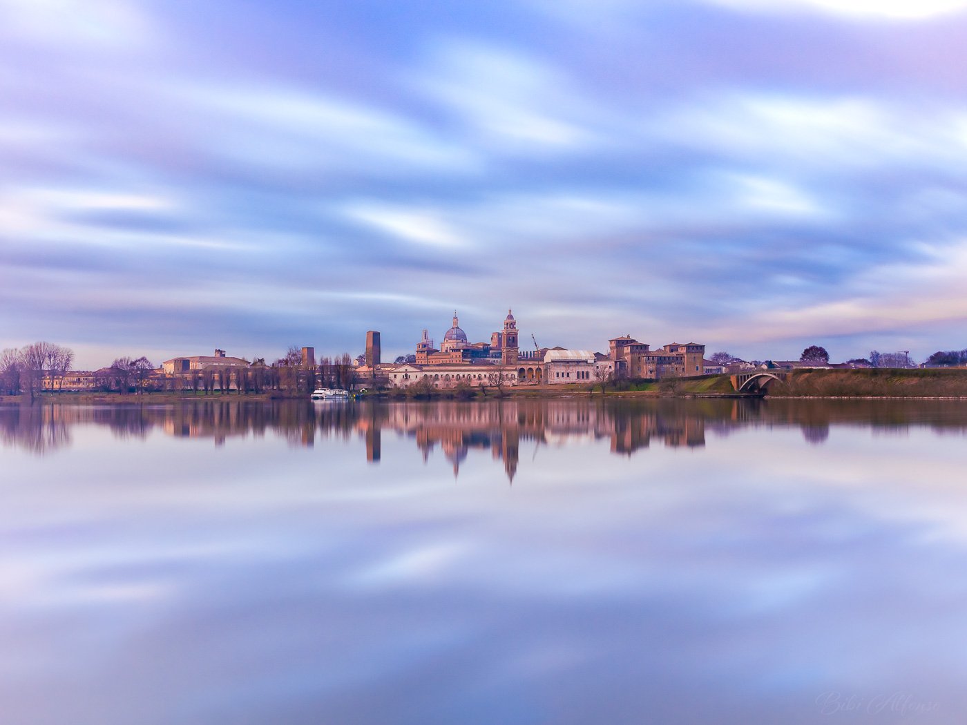 Spectacular long exposure of the Mantova skyline in the soft morning light, with streaky clouds above and the historic architecture reflected in the still water below, creating the effect of the city suspended in mid-air.