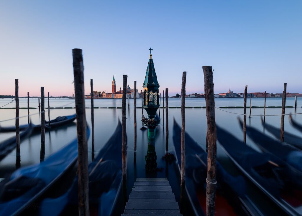 Long exposure shot at sunrise, with moving gondolas in the foreground and the Island of San Giorgio Maggiore in the background, capturing the motion and calm of Venice’s canals.