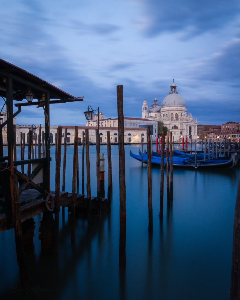 Long exposure blue hour photograph of Venice, Italy, featuring wooden mooring poles, gondolas on the lagoon, and the Basilica di Santa Maria della Salute illuminated in the background.