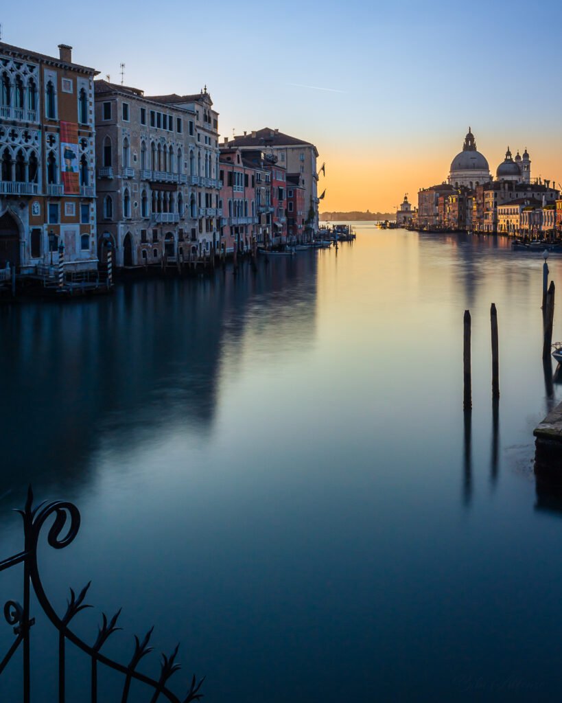 View of Punta della Dogana from Ponte dell’Accademia, with cold blue tones of the morning contrasting against the golden sunrise reflected on the dome of Santa Maria della Salute, captured in a long exposure shot.