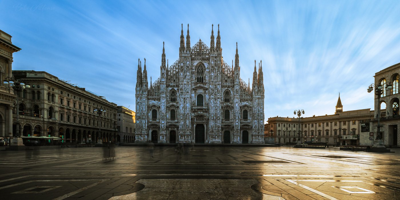Long exposure photo of Milan’s Duomo with blurred people and streaked clouds, capturing motion around the still cathedral.