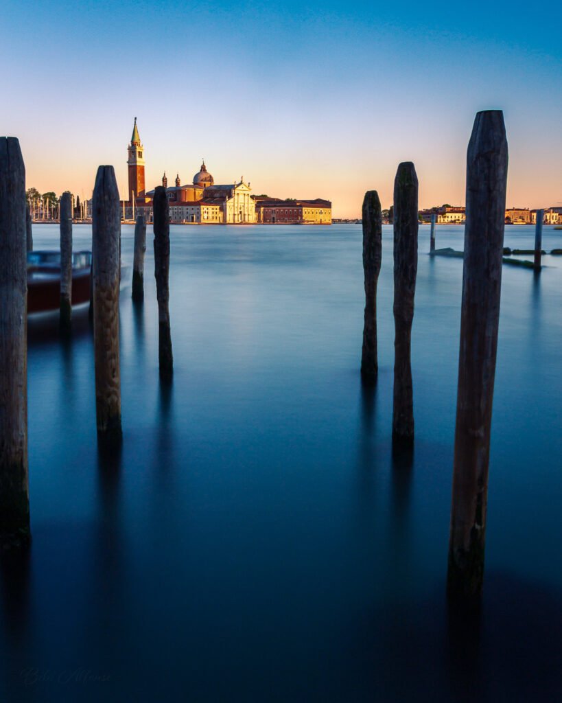 San Giorgio Maggiore illuminated by golden sunset light, with the sky transitioning from soft blue to warm golden hues, captured in a long exposure shot over the Venetian lagoon.