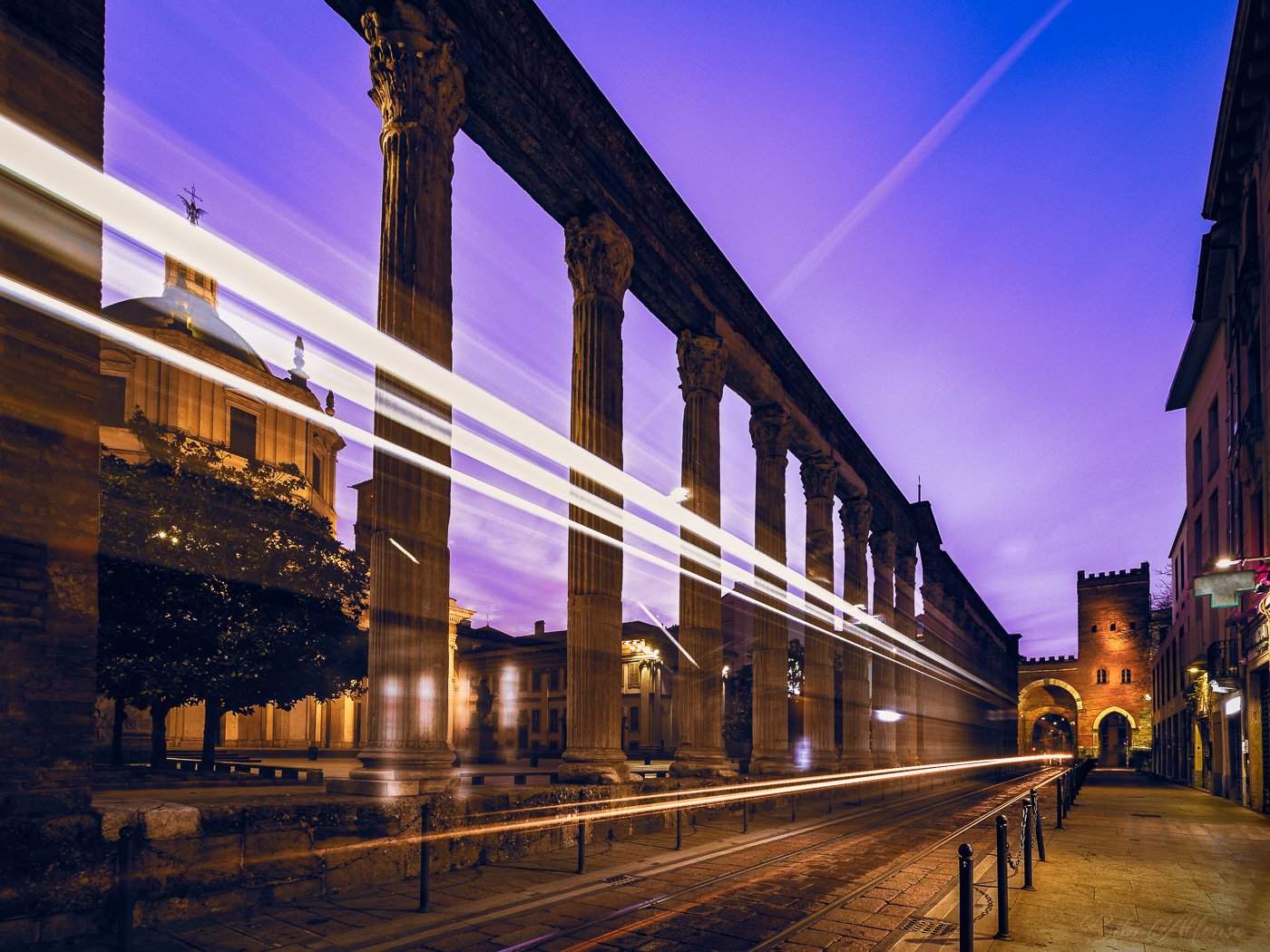 Very early morning long exposure at Colonne di San Lorenzo in Milan, with golden tram light trails weaving past the ancient Roman columns under a blue and purple sky.