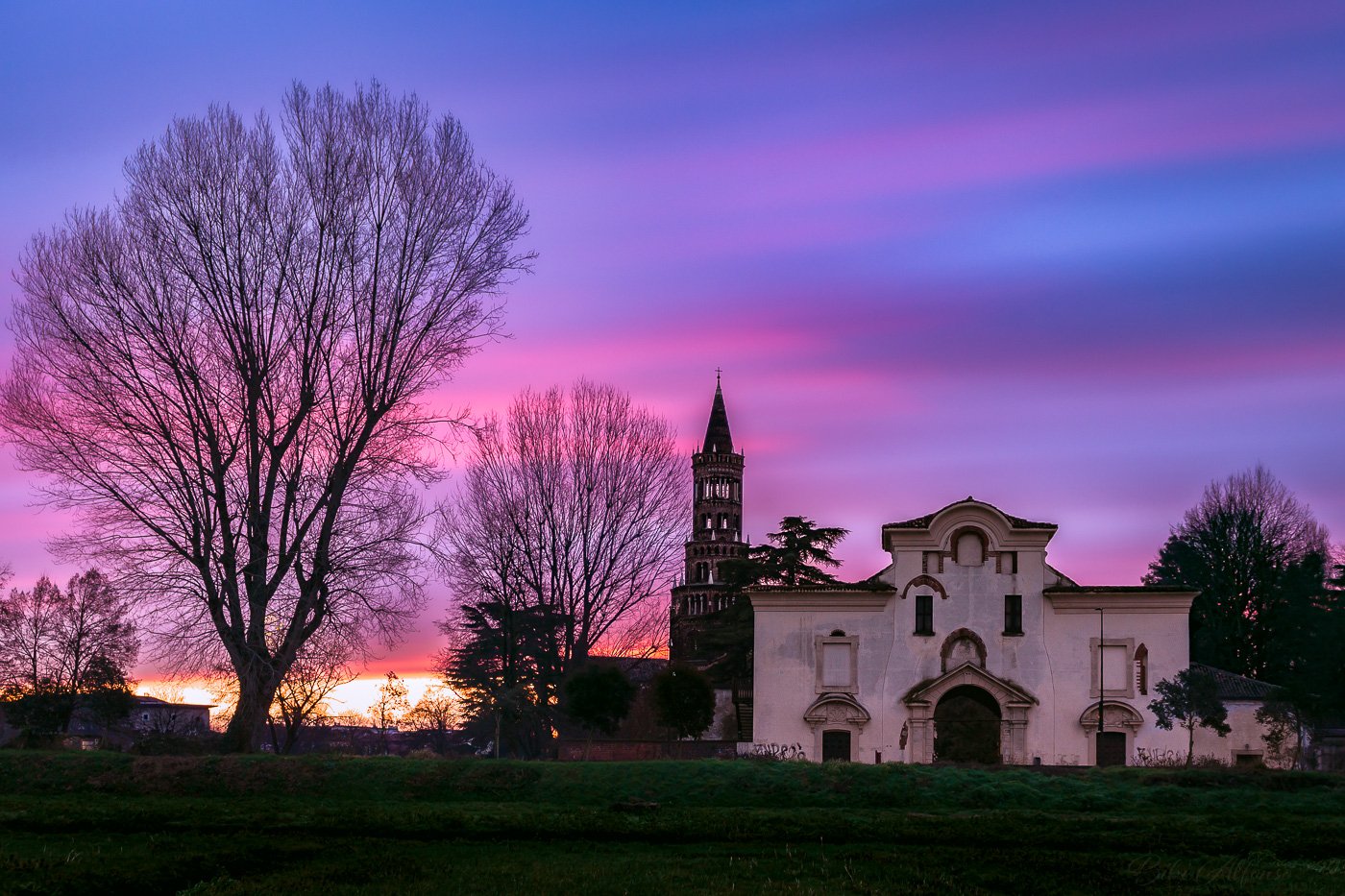 Long exposure winter sunrise over Chiaravalle Abbey south of Milan, Italy, featuring a church with a tall steeple surrounded by leafless trees under a purple, pink, and blue sky.