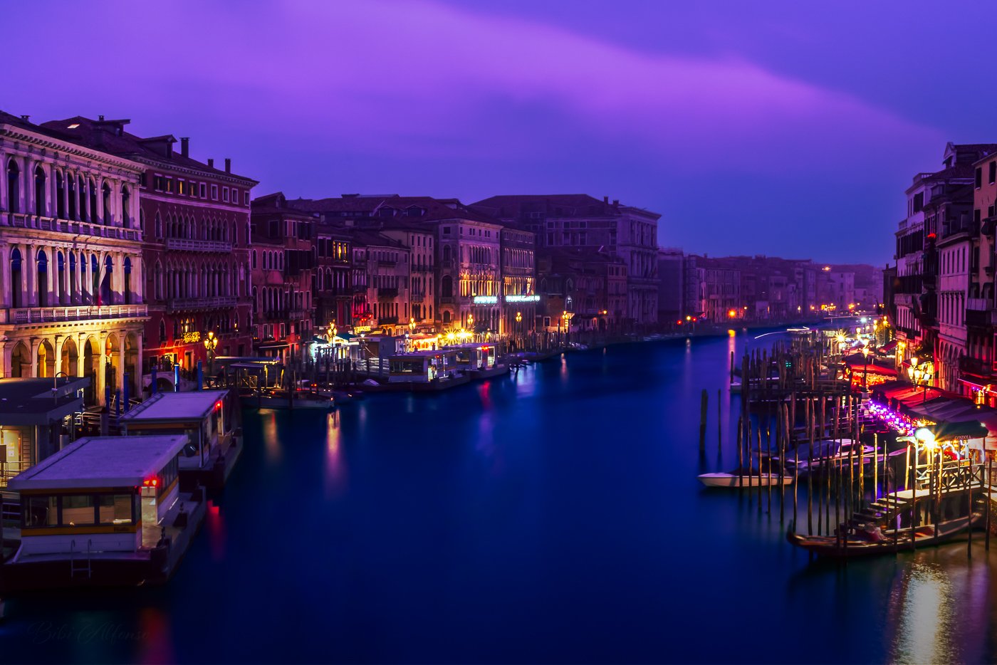 Long exposure twilight view of the Grand Canal from Rialto Bridge, Venice, Italy, featuring colorful historic buildings, illuminated shops, and boats docked along the water.
