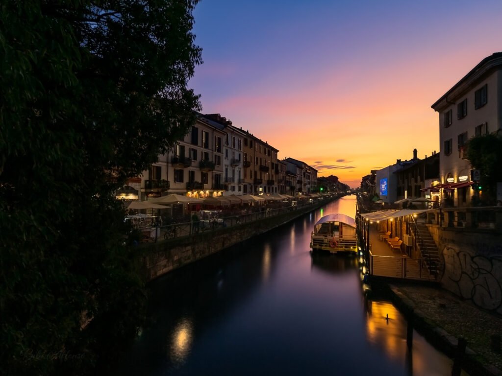 Long exposure twilight shot of Naviglio Grande canal in Milan, Italy, showing colorful buildings, outdoor cafes with umbrellas, boats, and streetlights glowing under sunset skies.