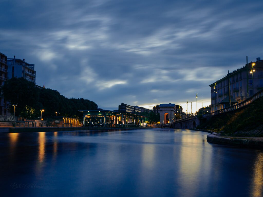 Long exposure at Darsena, Milan, transforming a windy morning into a serene scene, with calm waters reflecting golden lights from surrounding buildings under deep blue skies.