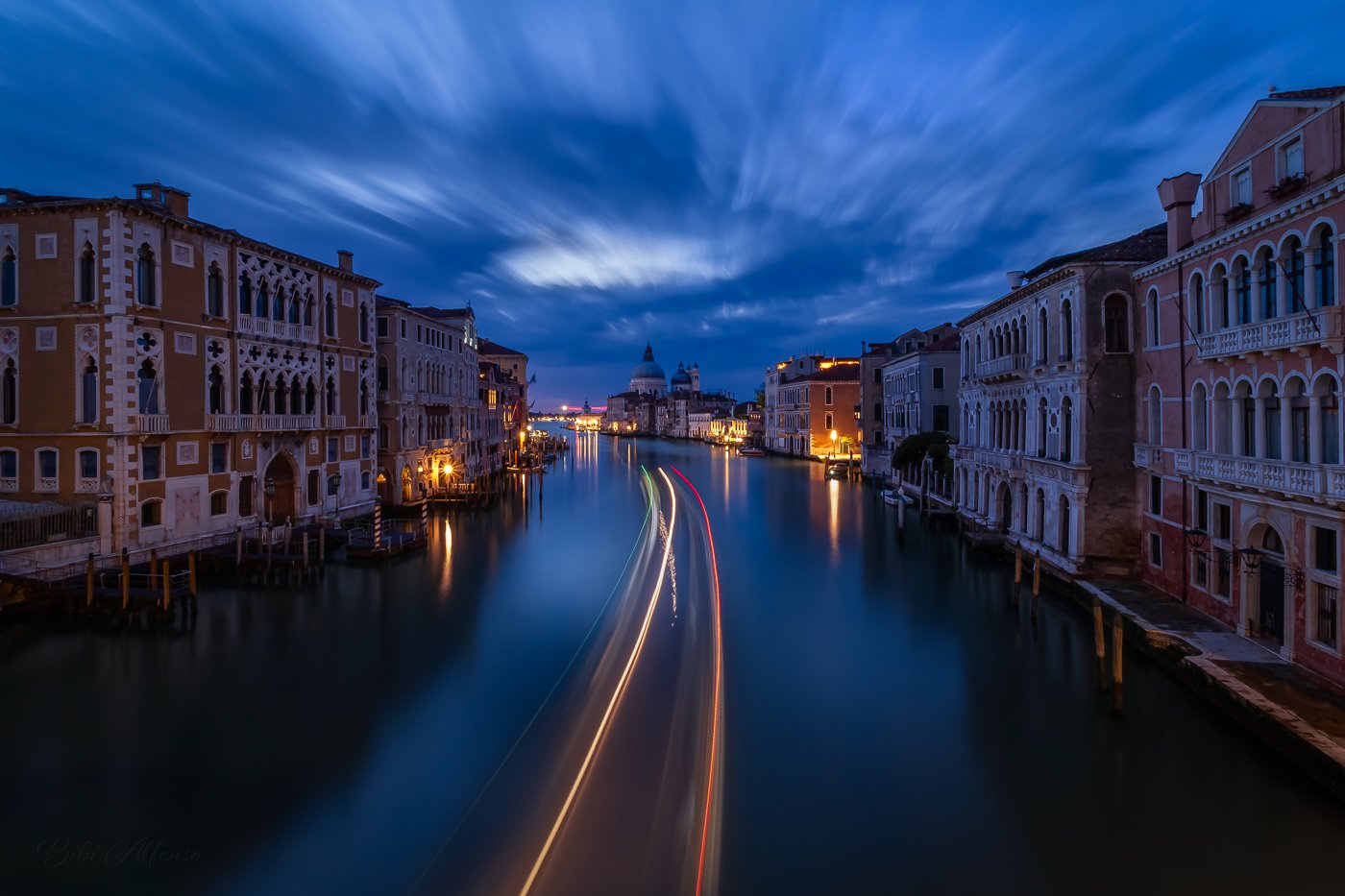 Venice Grand Canal at blue hour with long exposure light trails from boats leading toward Santa Maria della Salute, under streaked clouds and softly lit historic buildings on both sides.