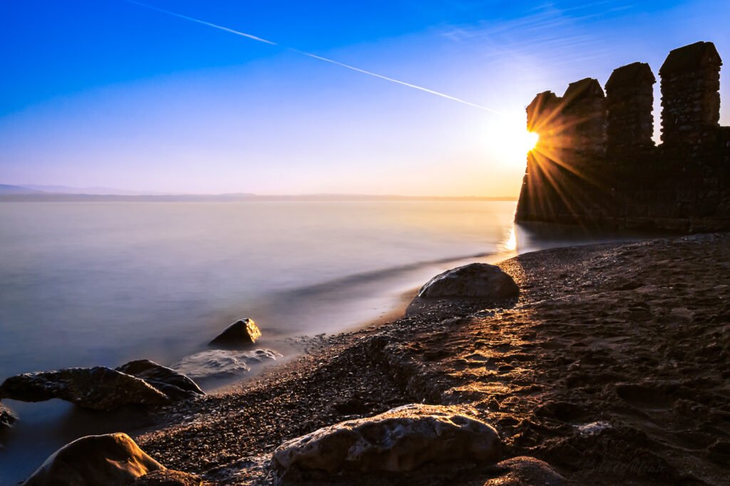 The sun rising behind the walls of Scaliger Castle in Sirmione, Lake Garda, creating a starburst effect in a long exposure shot with soft blue and golden tones.