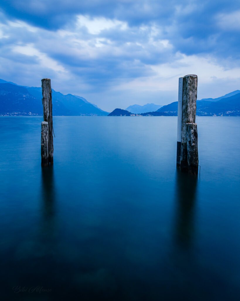 Long exposure view of Bellagio across Lake Como, framed in soft blue tones with calm water and distant shoreline dissolving into a tranquil horizon.
