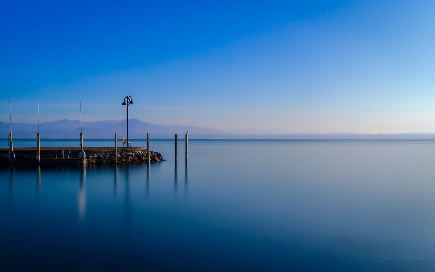Morning long exposure of Lake Garda at Sirmione, Italy, showing a calm lake with a small pier, bench, and lamppost under a clear blue sky, with distant mountains.