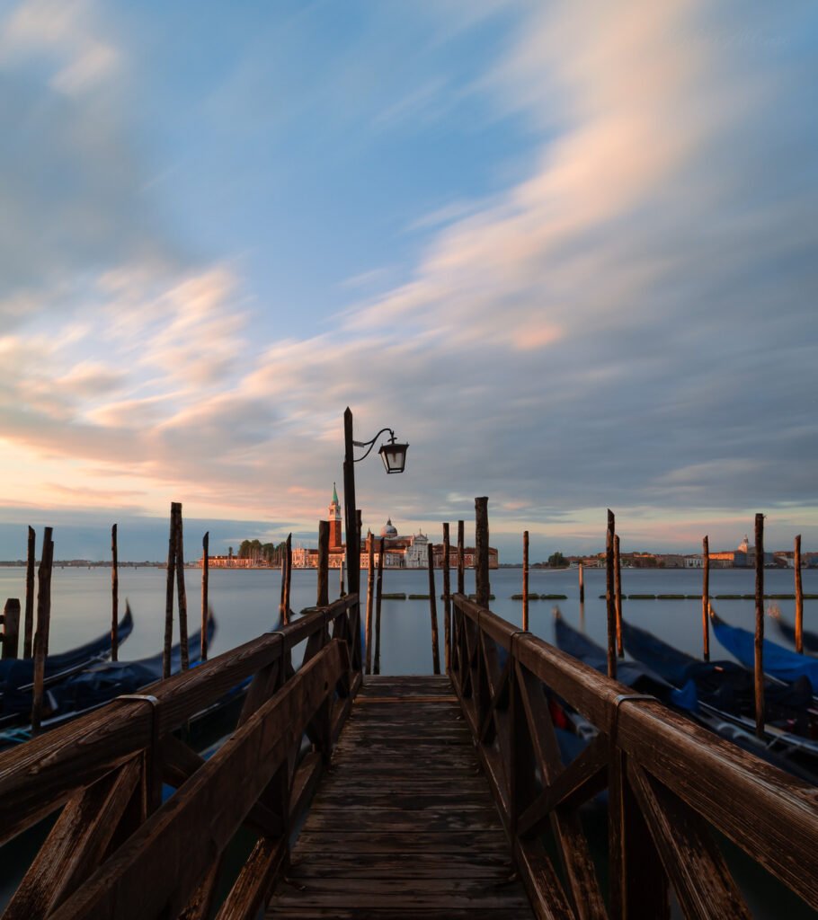Long exposure shot of San Giorgio Maggiore in Venice at sunrise, with a wooden pier in the foreground guiding the eye toward the island, blurred gondolas in the midground, and streaked clouds above. The scene glows with blues, browns, and golden tones.