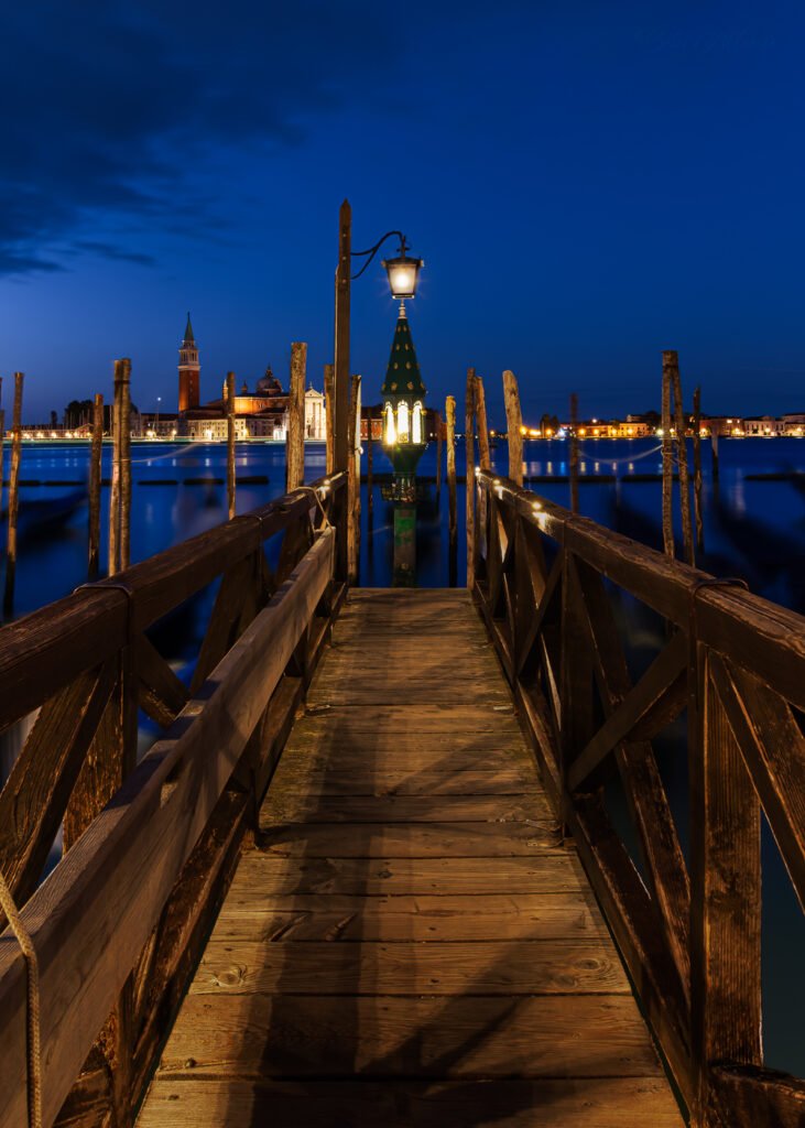 Long-exposure blue-hour photograph of a wooden Venetian pier leading toward a glowing lantern, with gondolas and wooden poles surrounding the calm lagoon and San Giorgio Maggiore illuminated in the distance under a deep cobalt sky.