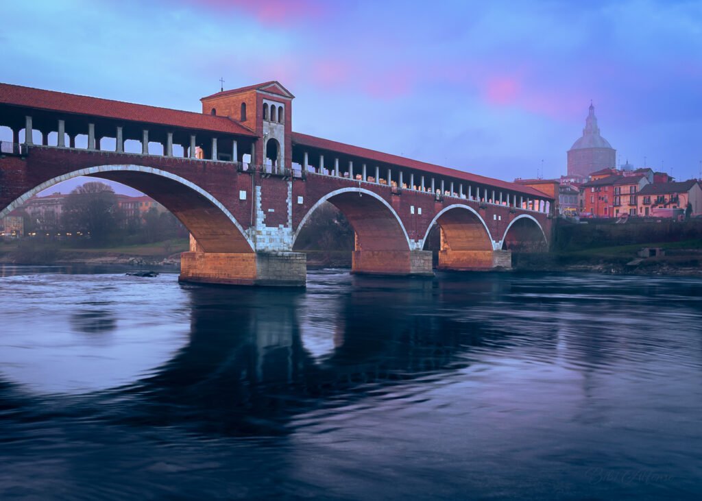 Foggy afternoon over Ponte Coperto in Pavia, captured in a fleeting moment of sunset light creating soft pink and blue hues, with the dark water of the Ticino River below.