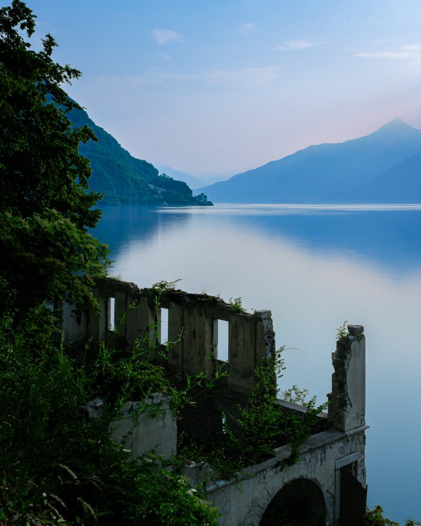 Long exposure sunrise in Menaggio on Lake Como, Italy, featuring the ruins of an abandoned building in the foreground, calm smoothed water in the midground, and sunlit mountains with green vegetation in the background, captured in a serene travel photography scene.