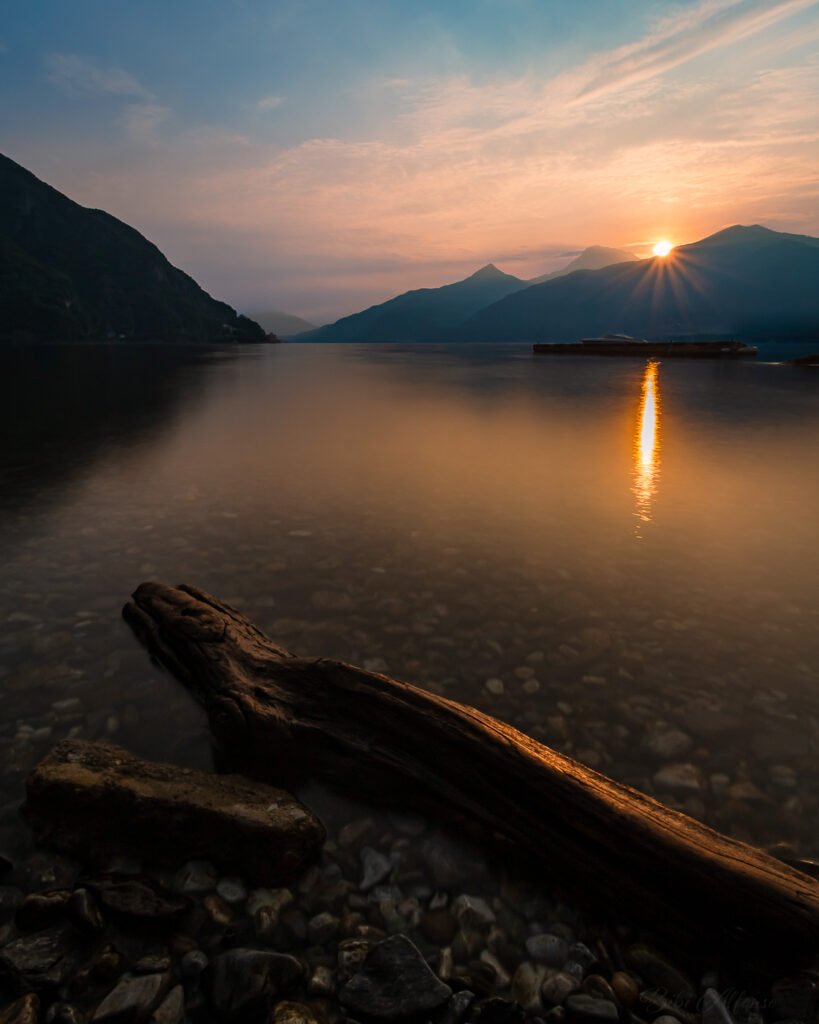 Long exposure of sunrise in Menaggio, Lake Como, featuring a log in the foreground, sunburst behind mountain silhouettes, and warm orange light spreading across the water under a soft blue morning sky.