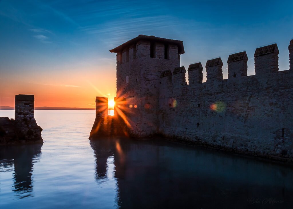 Dramatic sunrise behind the walls of Scaliger Castle in Sirmione, Lake Garda, with a starburst effect over the fortifications and warm golden light contrasting the cool blue water.