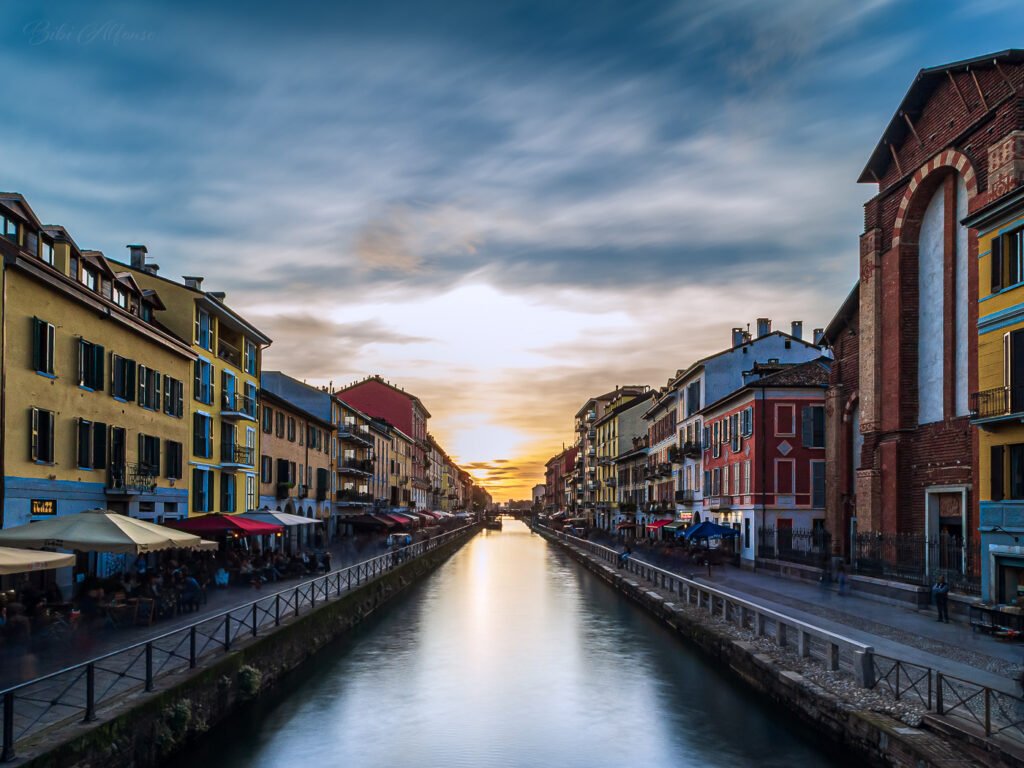 30-second long exposure of a sunset over Naviglio Grande, Milan, featuring silky water reflecting blue and yellow tones, with surrounding red brick buildings adding contrast.