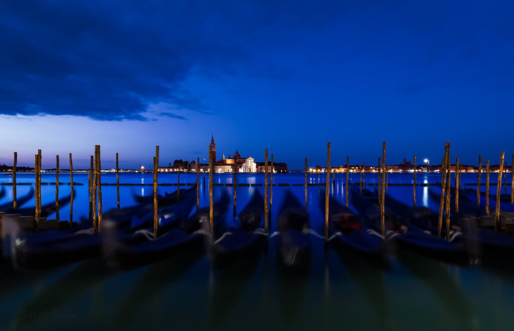 Long-exposure blue-hour photograph of gondolas and wooden mooring poles facing San Giorgio Maggiore across the Venetian lagoon, with deep cobalt sky reflections and soft motion blur in the foreground boats.