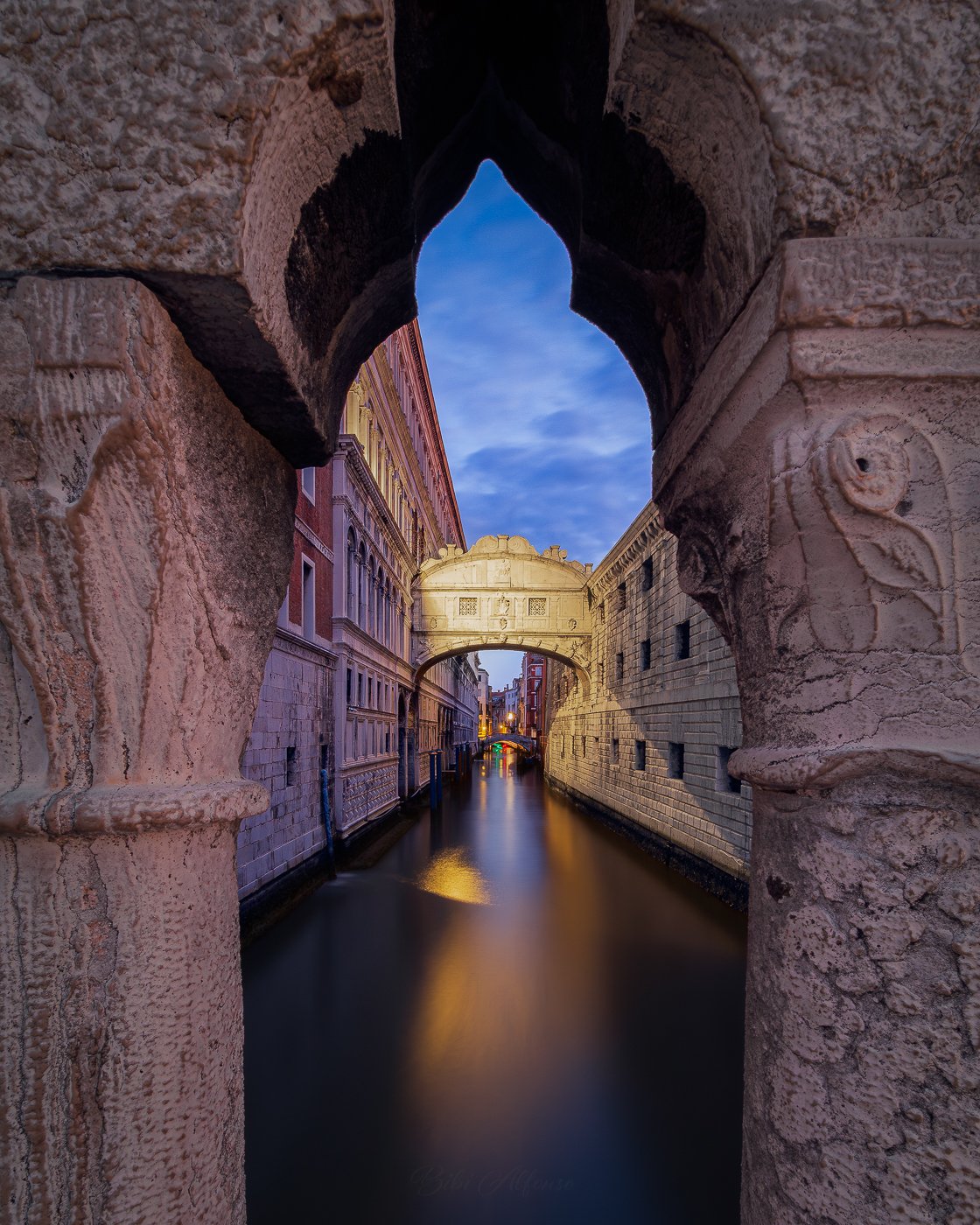 Long exposure photograph of the Bridge of Sighs in Venice at blue hour, framed through a stone Gothic opening above a calm canal with warm reflections and soft morning light.