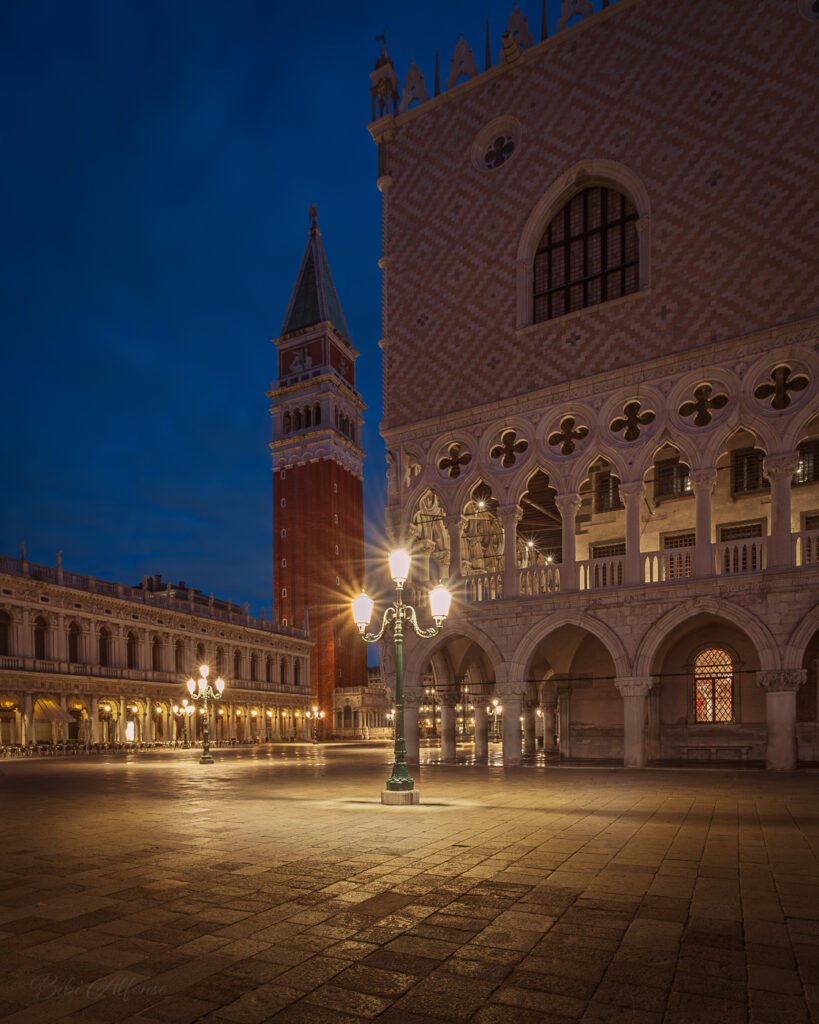 Venice at blue hour near St Mark’s Square, with the Doge’s Palace in the foreground and St Mark’s Campanile rising in the background, lit by warm street lamps over an empty stone square beneath a deep blue sky.