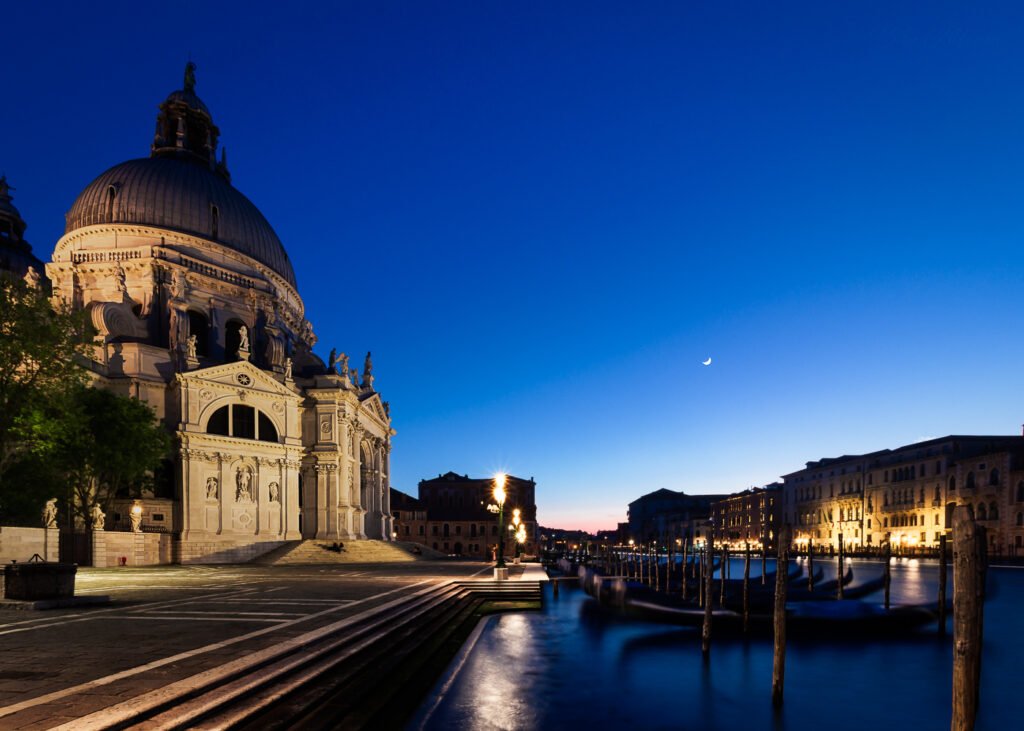 Long exposure after-sunset shot of the Grand Canal in Venice, featuring Campo della Salute and Santa Maria della Salute, with gondolas docked along the canal and golden lights reflecting on deep blue waters.