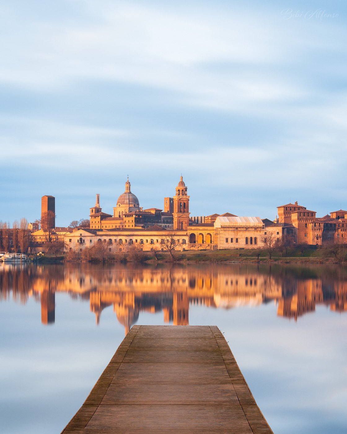 Long-exposure photograph of the skyline of Mantova reflected in calm water on a winter morning, with a wooden pier leading toward historic buildings illuminated by warm sunrise light beneath a soft blue sky.
